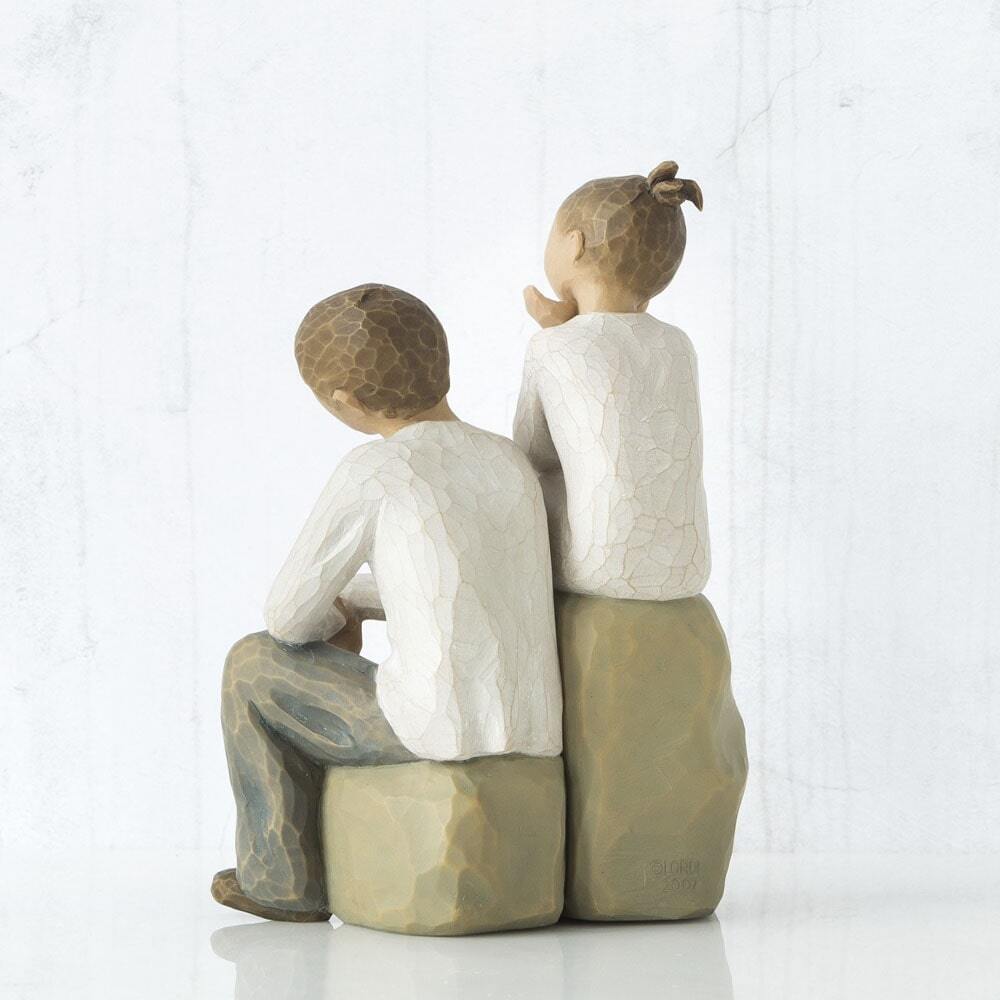 Figure of boy and girl, seated on a gray rock next to each other. Girl in cream dress, boy in cream shirt and blue jeans