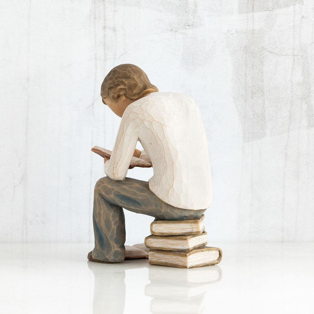 Figure of young man in cream shirt and blue jeans, seated on stack of books, reading book