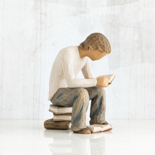 Figure of young man in cream shirt and blue jeans, seated on stack of books, reading book