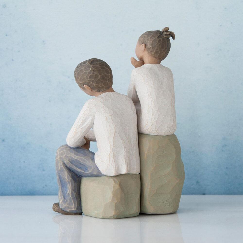 Figure of boy and girl, seated on a gray rock next to each other. Girl in cream dress, boy in cream shirt and blue jeans