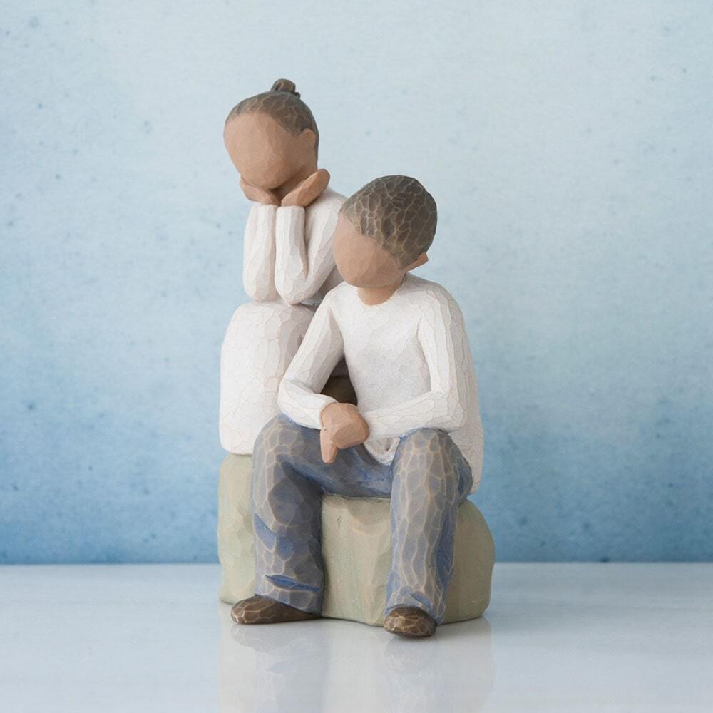 Figure of boy and girl, seated on a gray rock next to each other. Girl in cream dress, boy in cream shirt and blue jeans